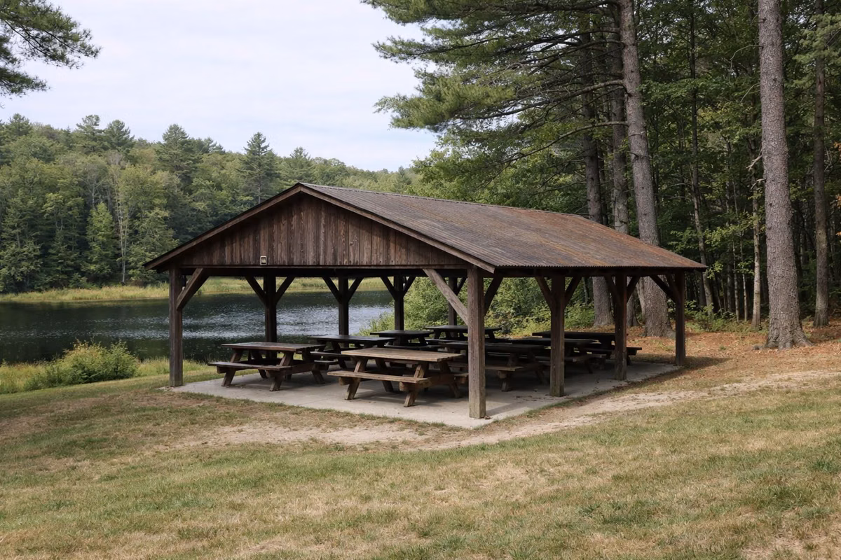 An open timber picnic pavilion on a grassy slope near a pond